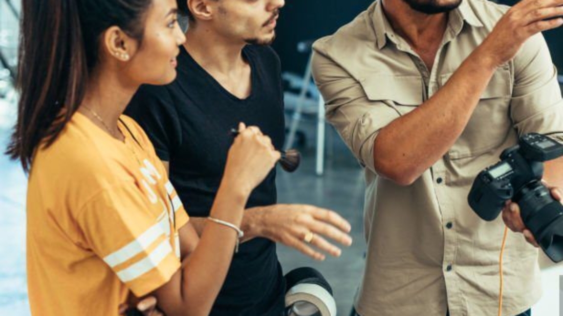 Photographer explaining about the shot to his team in the studio. Photographer talking to his assistants holding a camera during a photo shoot.