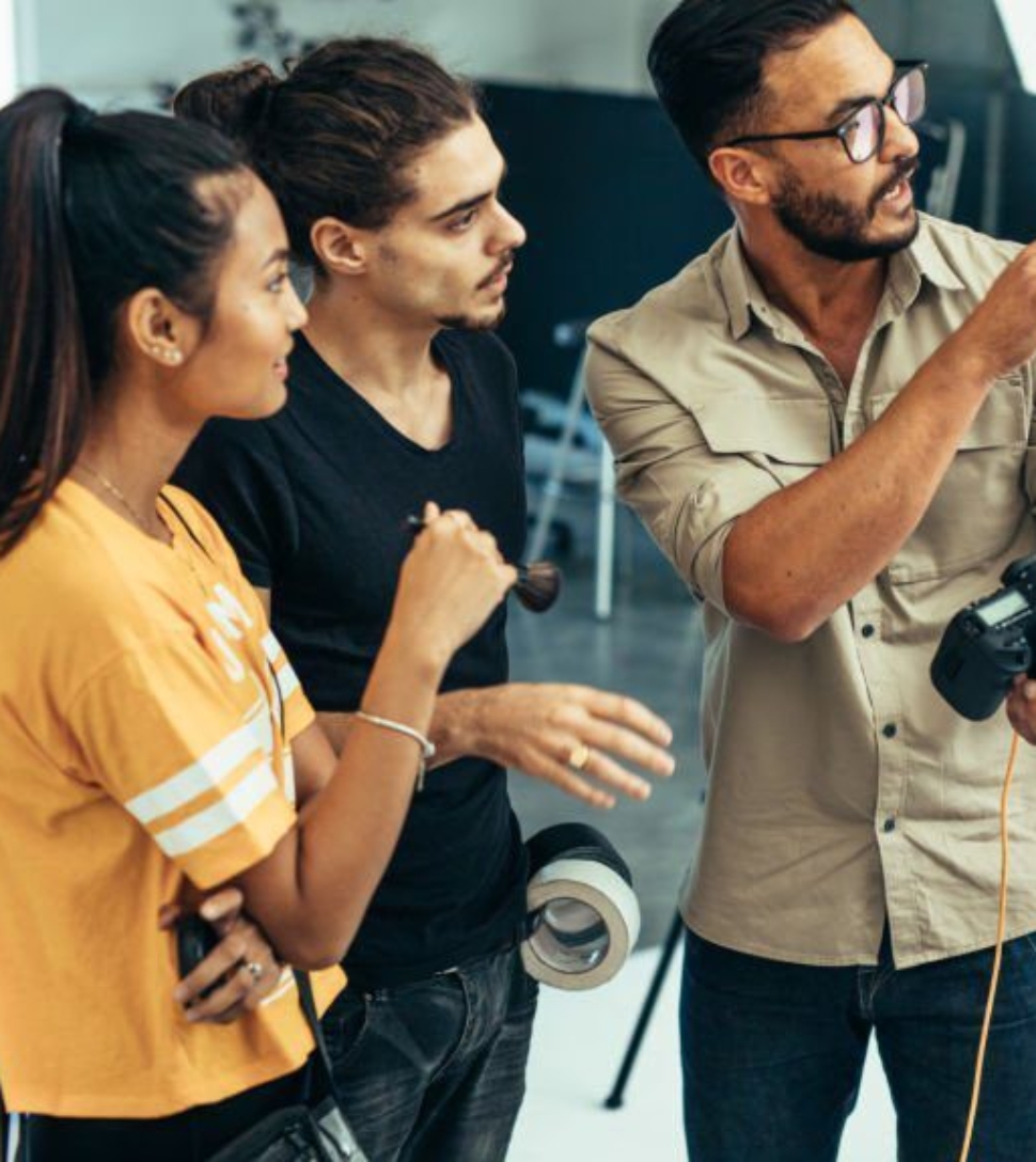 Photographer explaining about the shot to his team in the studio. Photographer talking to his assistants holding a camera during a photo shoot.